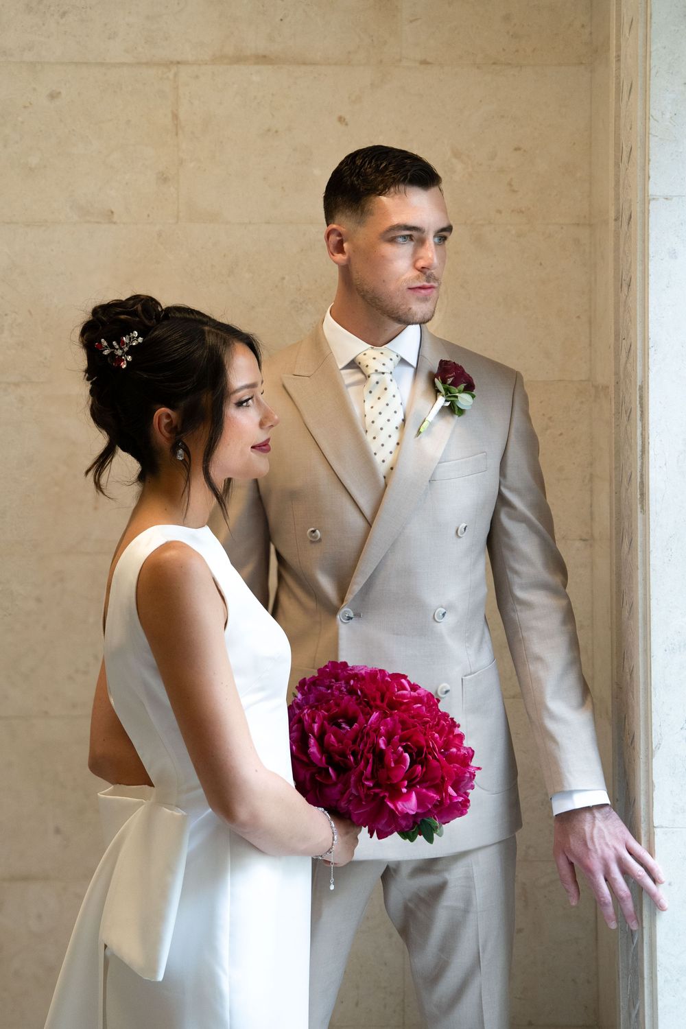 A bride in a white dress holds vibrant magenta peonies while posing with someone in a light gray suit at their wedding.