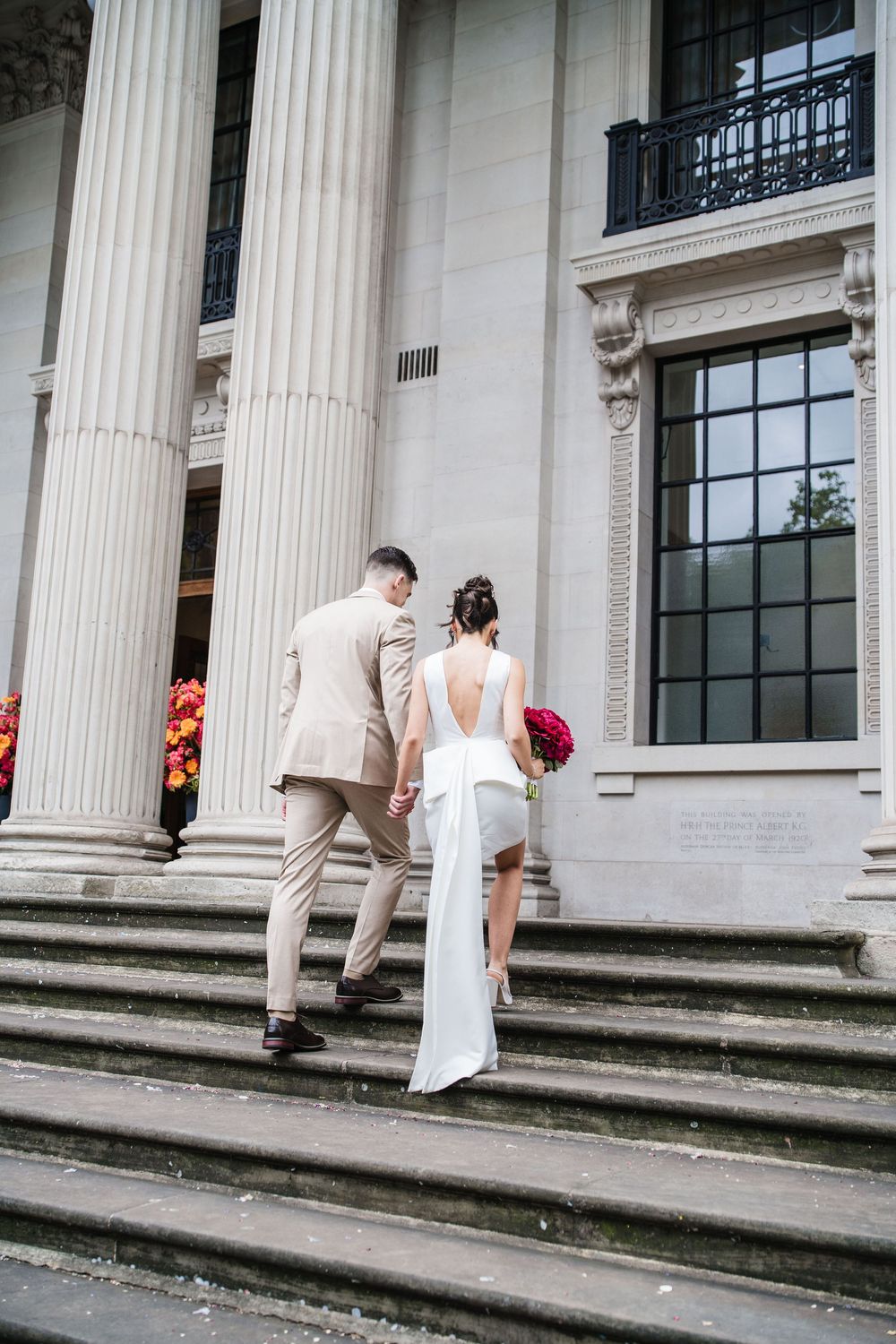 Newlywed couple ascends stone steps of a grand columned building in formal wedding attire and bouquet.