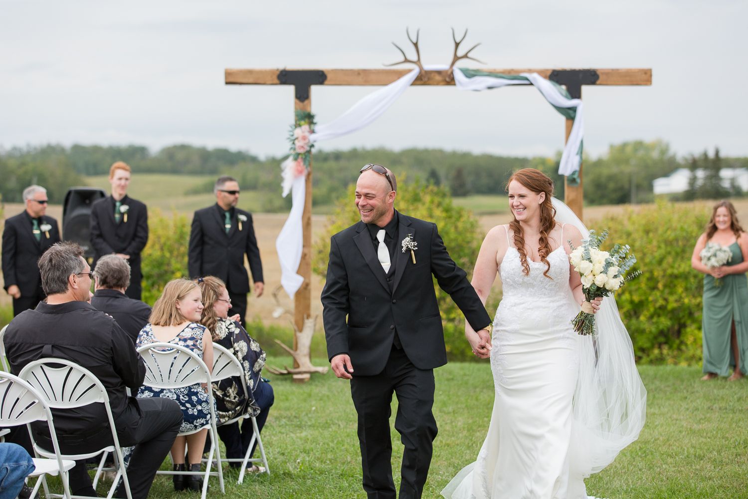 Joyful newlyweds walking down aisle after outdoor ceremony under rustic wooden arch.