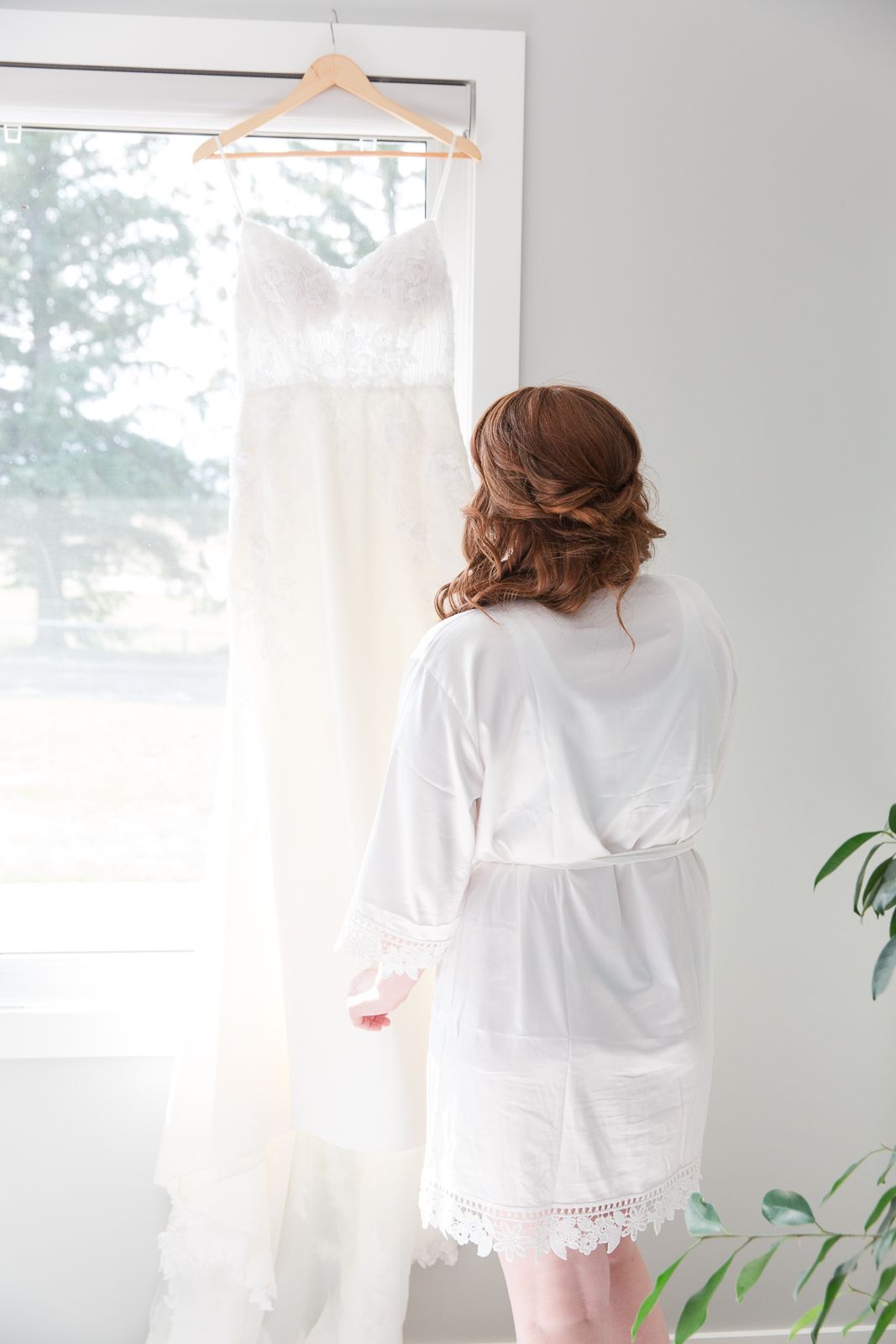 A white lace wedding dress hangs by a window as someone admires it.