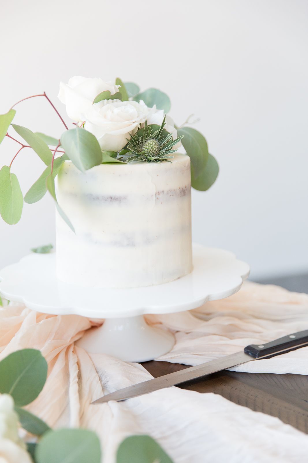 A delicate white wedding cake decorated with eucalyptus leaves on a white pedestal.
