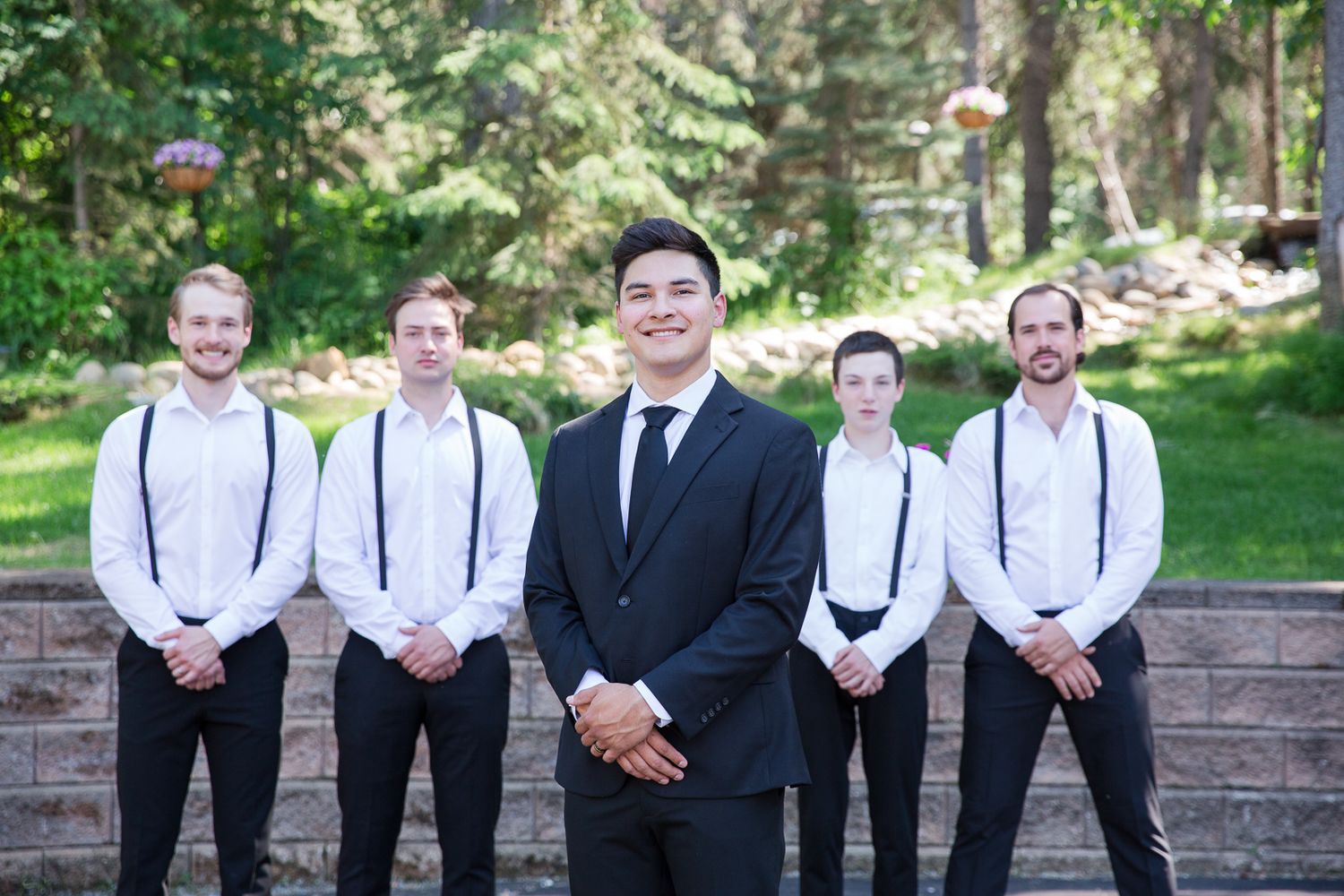 Groomsmen in suspenders and black suits pose together in garden setting.