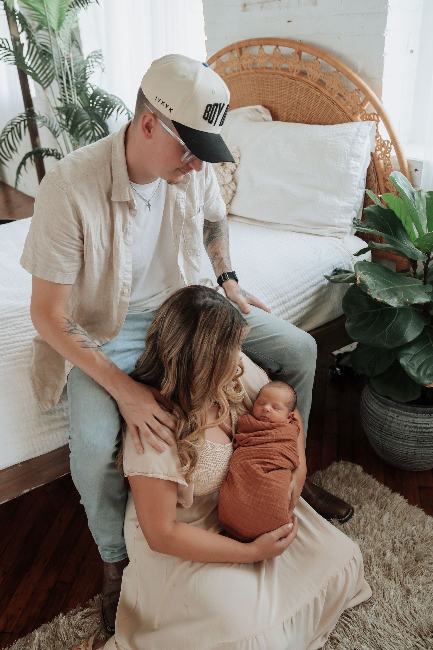New parents lovingly admire their newborn baby wrapped in a rust-colored swaddle in a cozy bedroom.