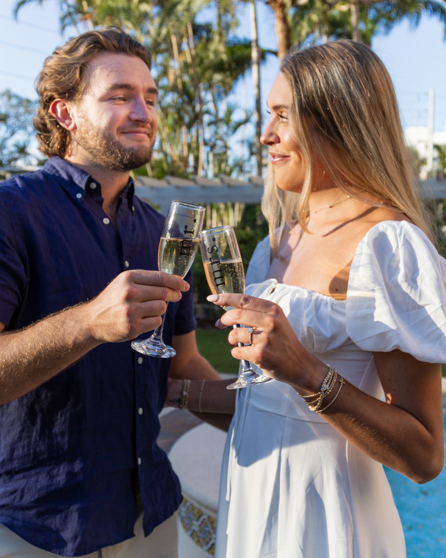 Happy couple toasting with champagne glasses outdoors near a pool at golden hour.