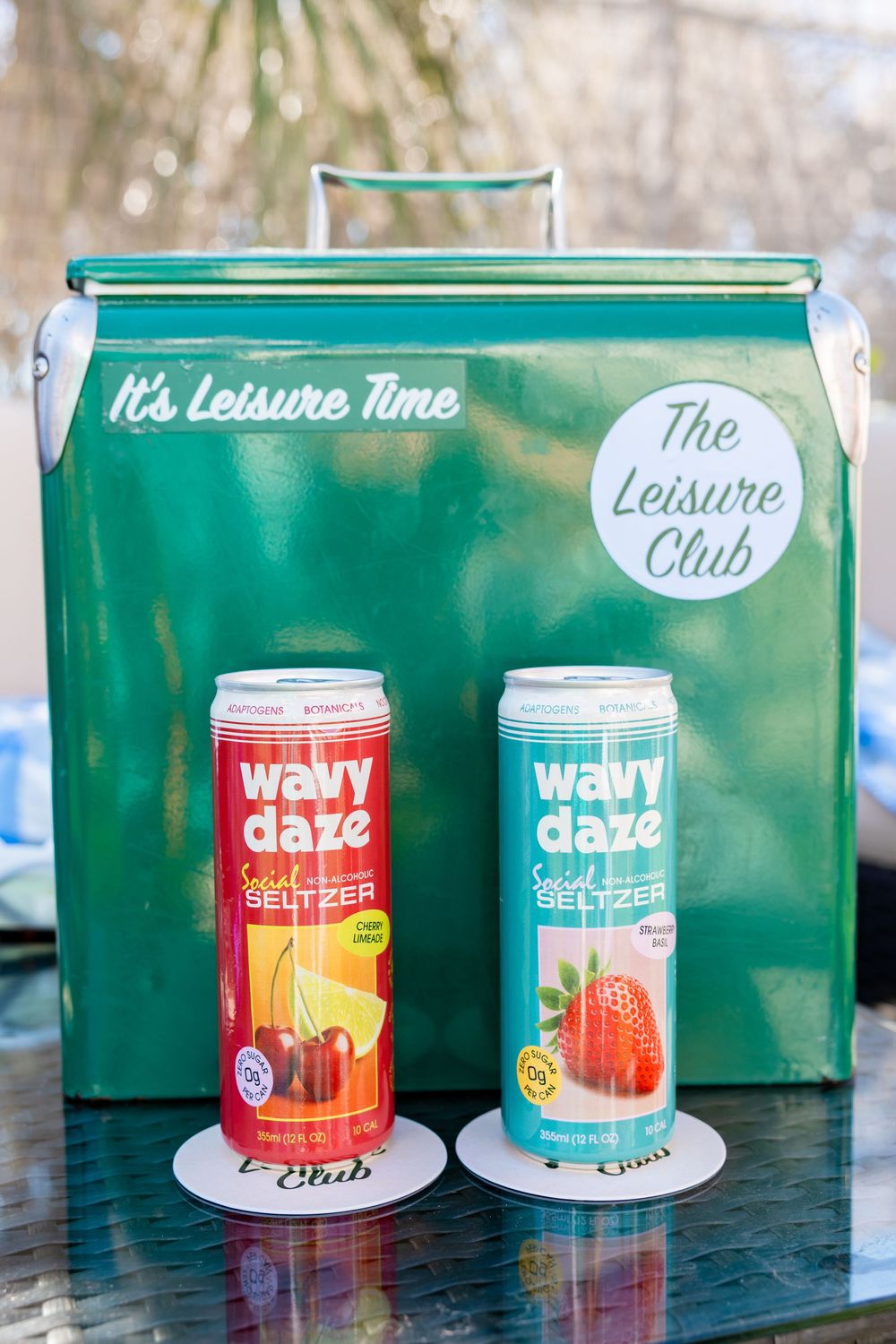 Two Wavy Daze non-alcoholic seltzer cans in front of green Leisure Club vintage cooler.