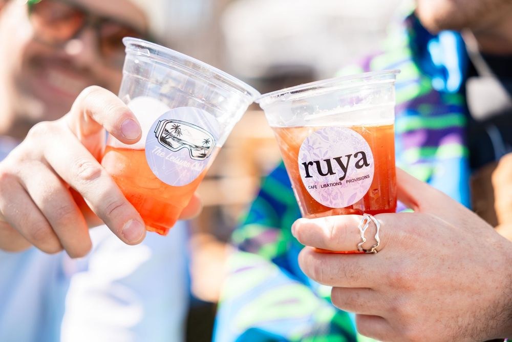 Two people clinking Ruya branded plastic cups with orange cocktails outdoors.