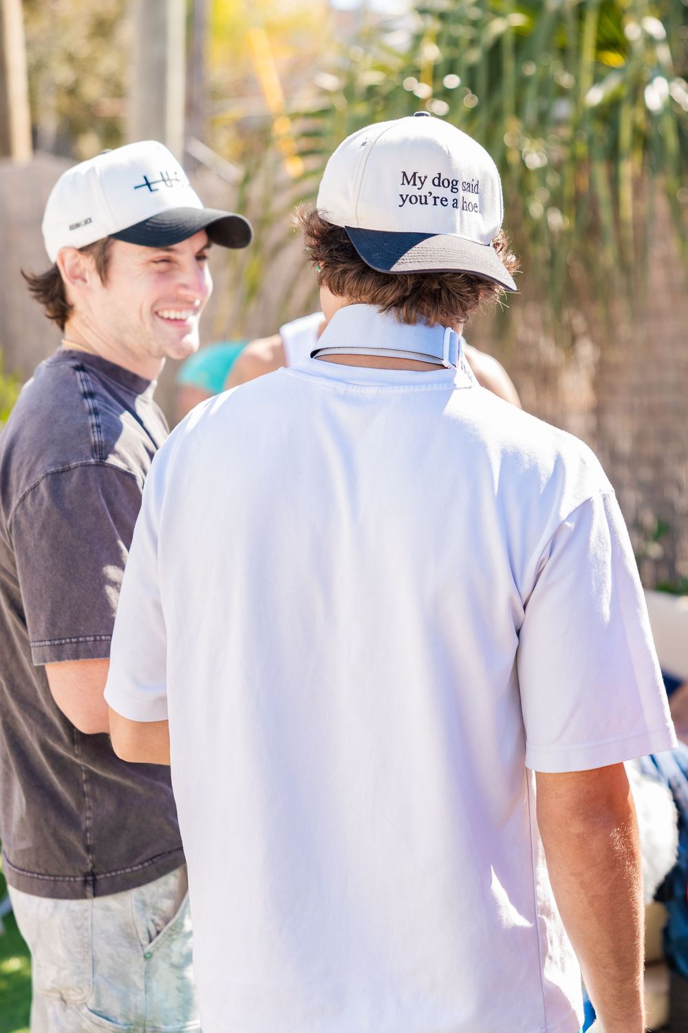 Two young men wearing white baseball caps chatting at an outdoor social gathering.