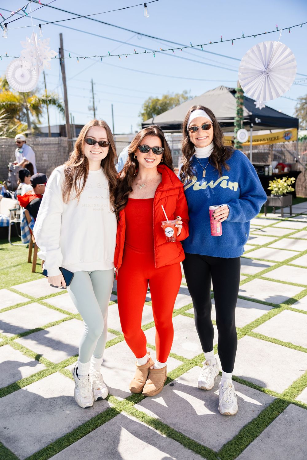 Three women in casual winter outfits posing together at a sunny outdoor event.