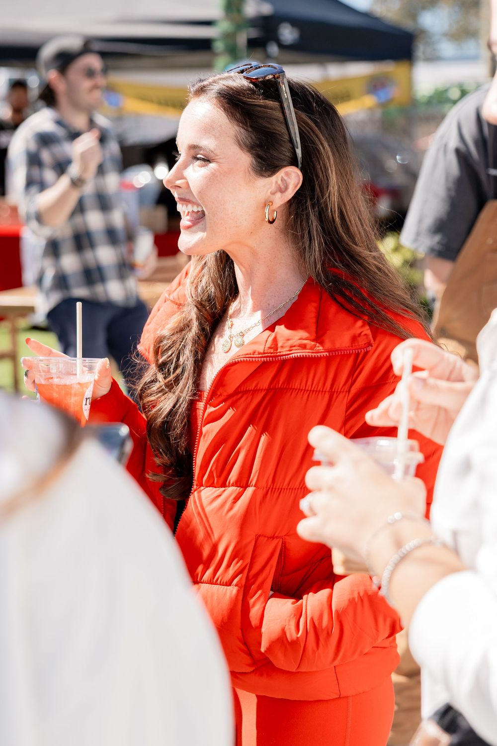 Laughing woman in red outfit holding a drink at a sunny outdoor gathering.