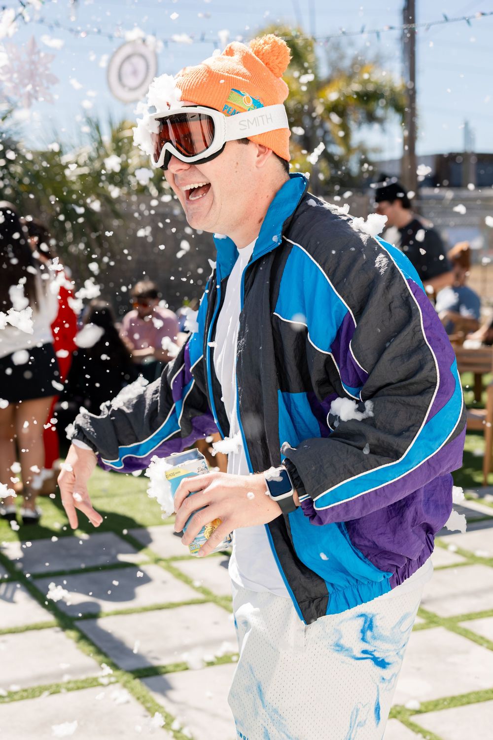 Man in retro ski jacket and orange beanie laughing in artificial snow at party.