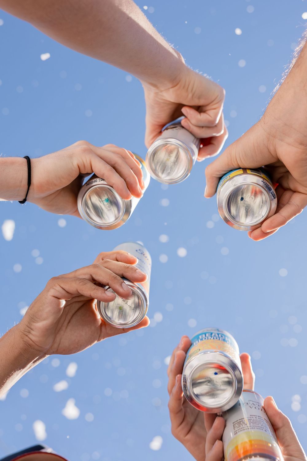 Four hands toasting with canned drinks against a blue sky with snow falling.