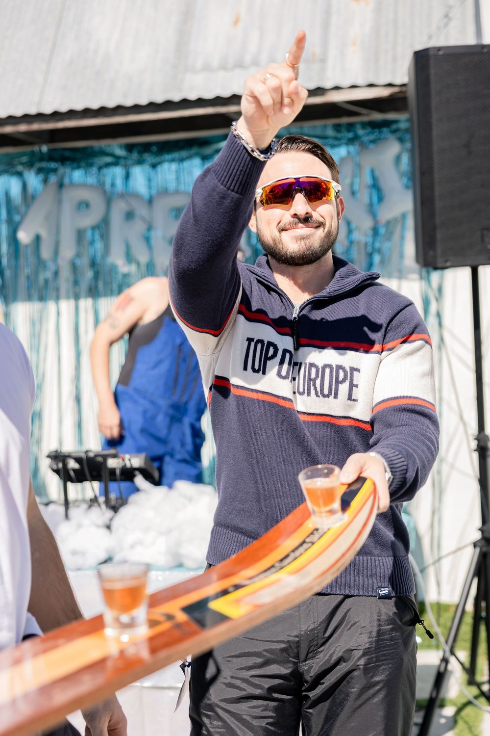 Bearded man in Top of Europe sweater pointing up while holding ski shot board.