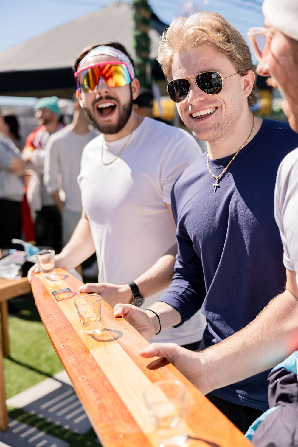 Two smiling men in sunglasses playing a drinking game at a sunny outdoor event.