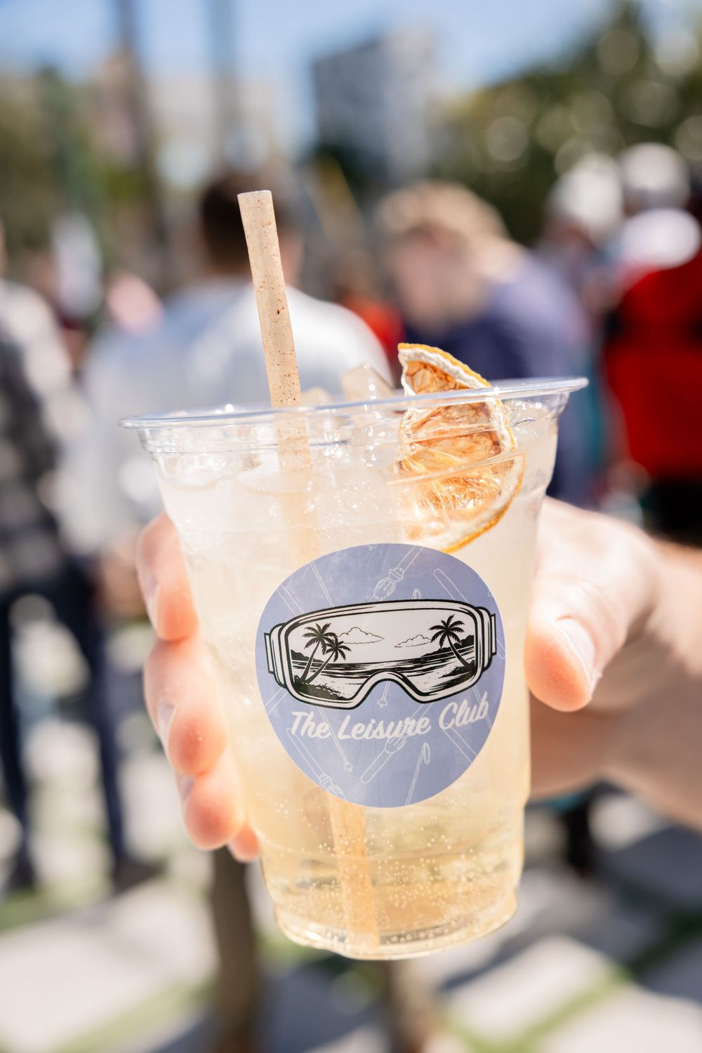 Hand holding a The Leisure Club branded cocktail cup with citrus garnish and paper straw at outdoor event.