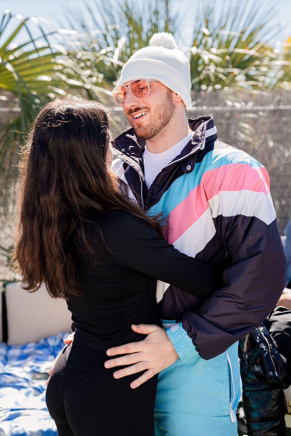 Couple embracing outdoors, man wearing colorful retro ski jacket, white beanie, and pink sunglasses.
