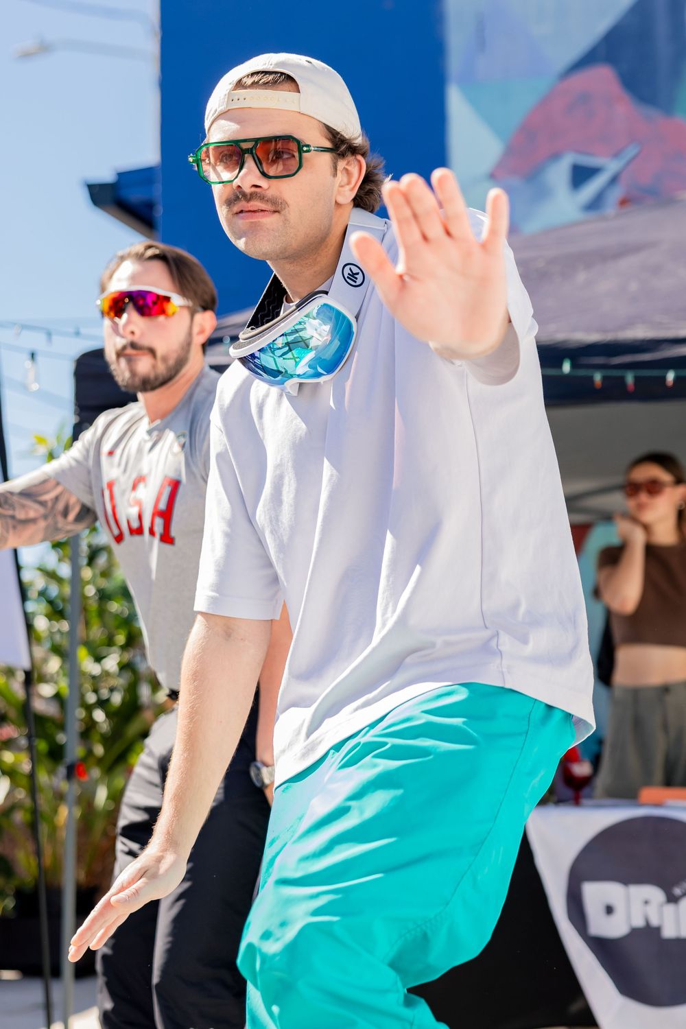 Man in white tee and teal shorts with headphones gesturing on outdoor stage at sunny festival.