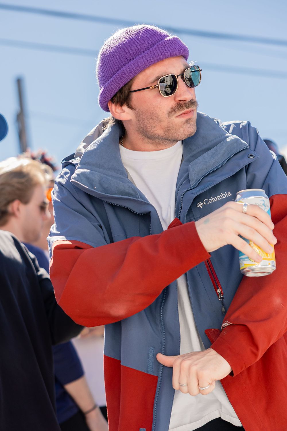 Man wearing purple beanie, Columbia jacket, and sunglasses holding a canned drink at outdoor event.