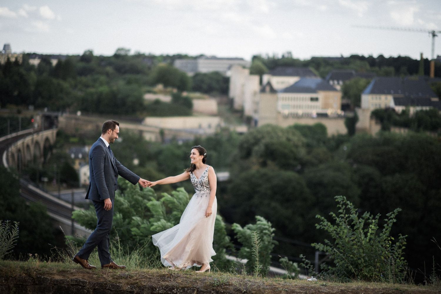 A couple in wedding attire holding hands while walking along a hilltop path with buildings in the distance.