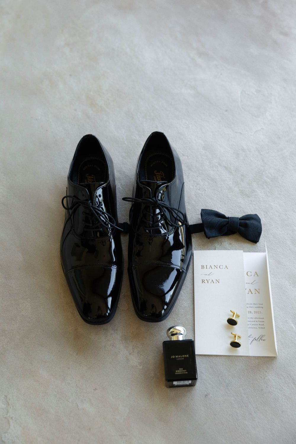 Black patent leather dress shoes with cufflinks and wedding invitation on white background.