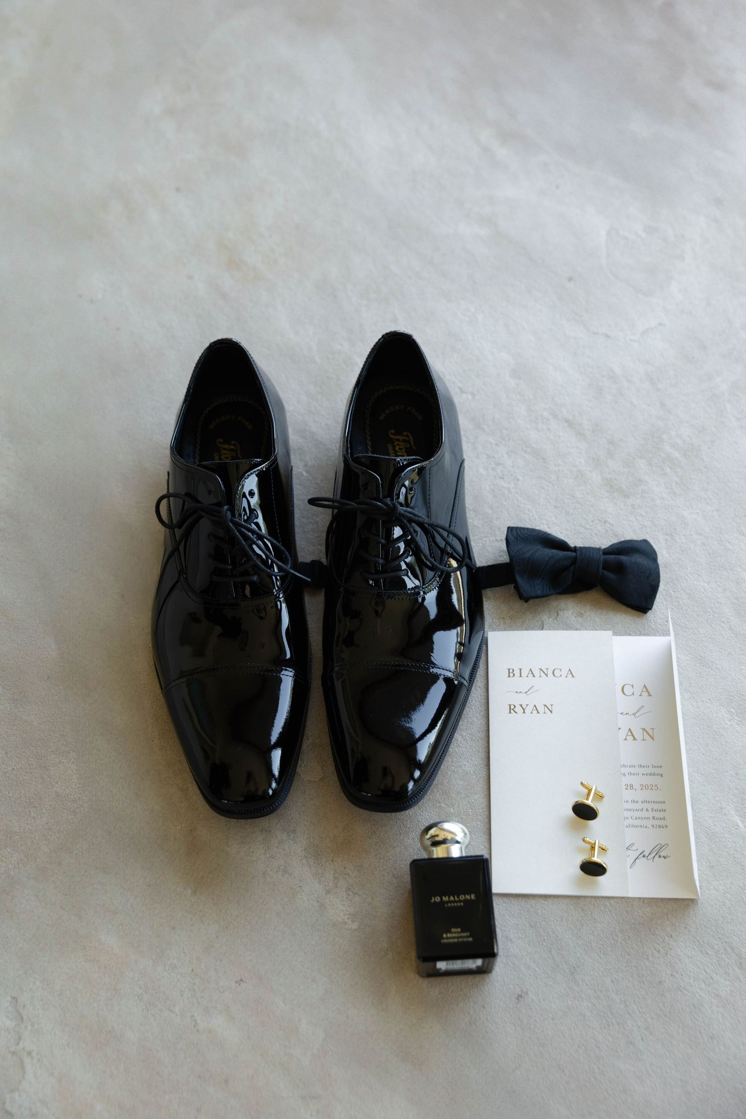 Black patent leather dress shoes with cufflinks and wedding invitation on white background.