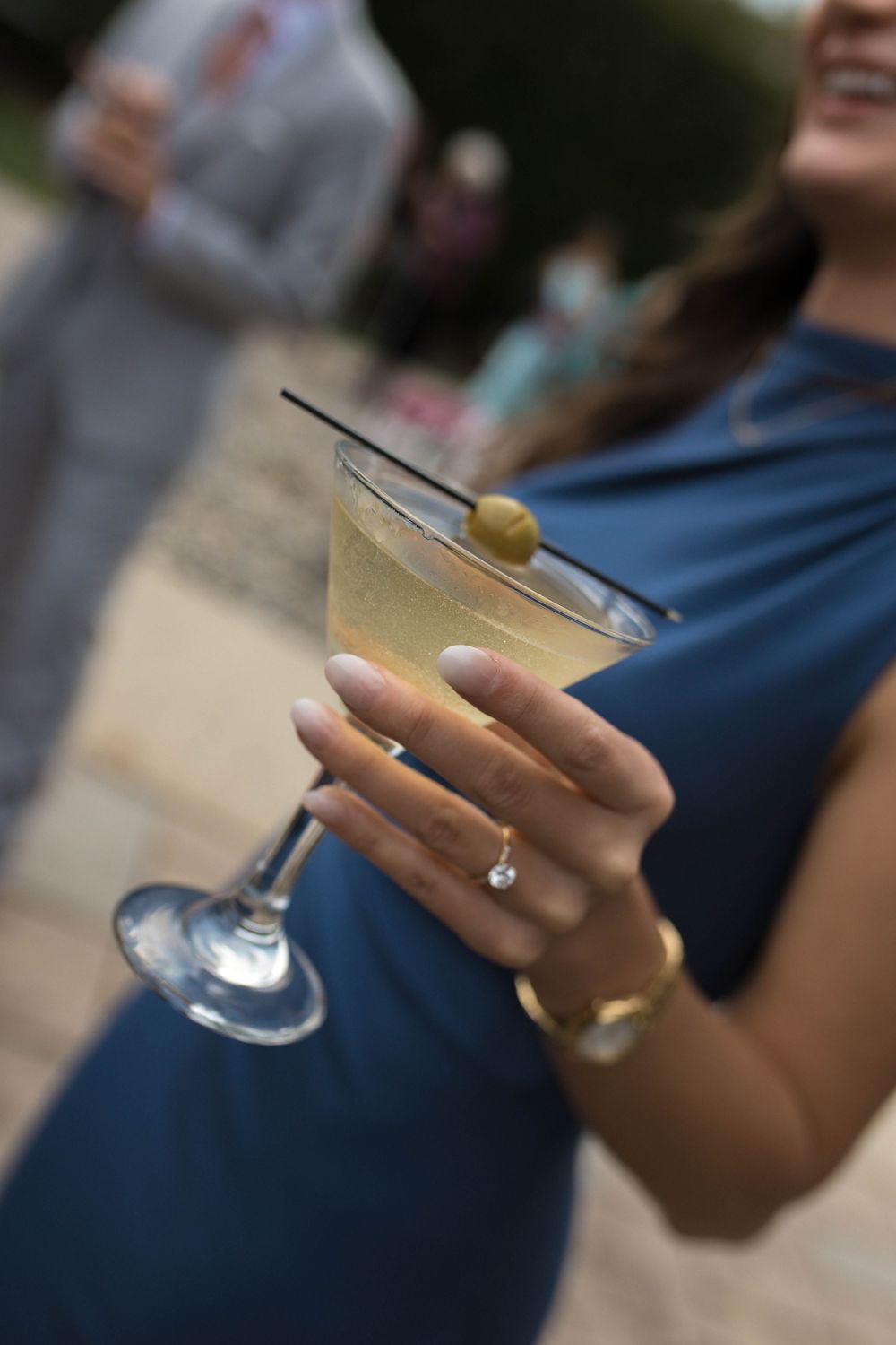 A hand holding a martini glass with an olive garnish against a blurred background.