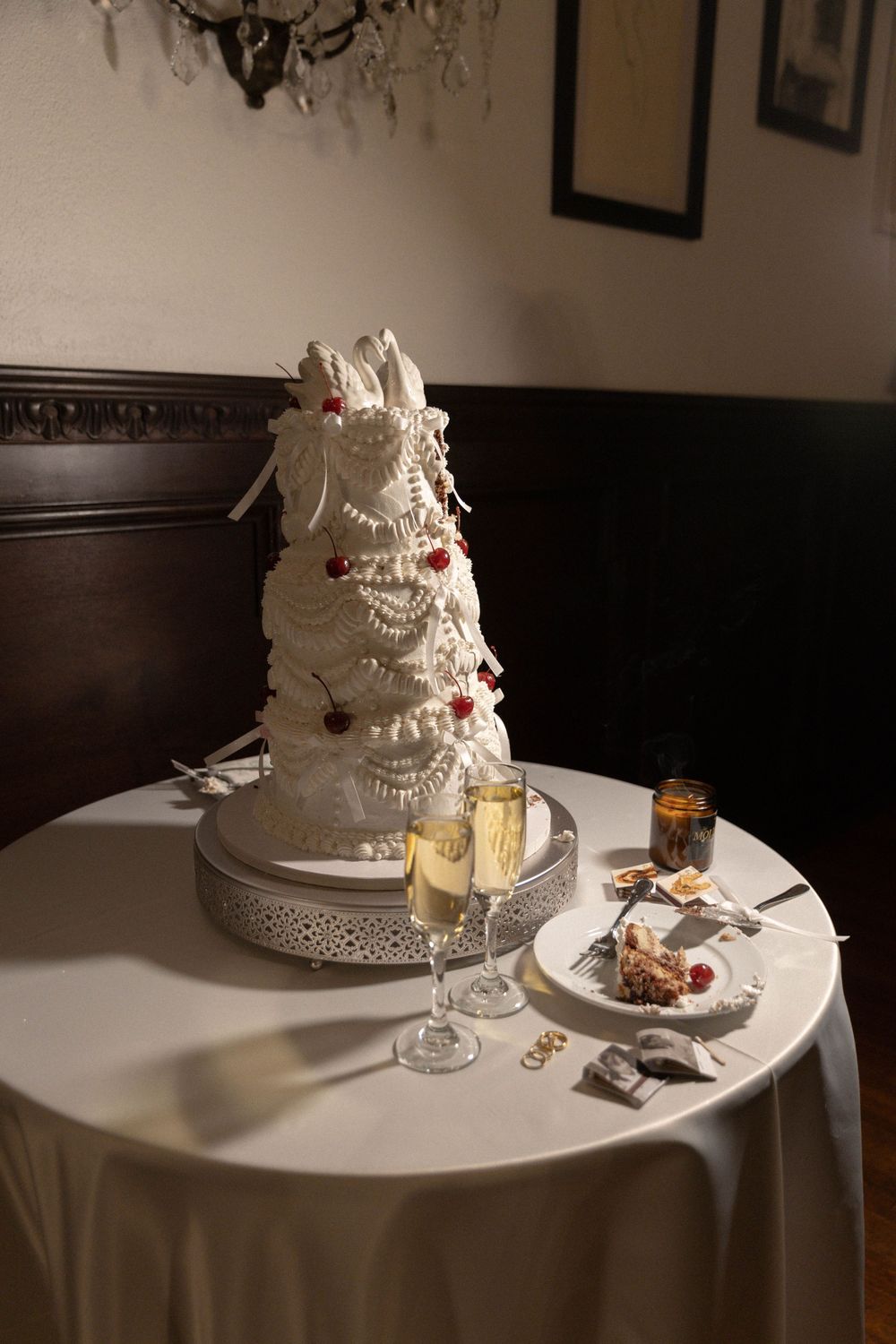 An elegant white wedding cake with intricate lace details and pearl accents sits on a round table with a white tablecloth.