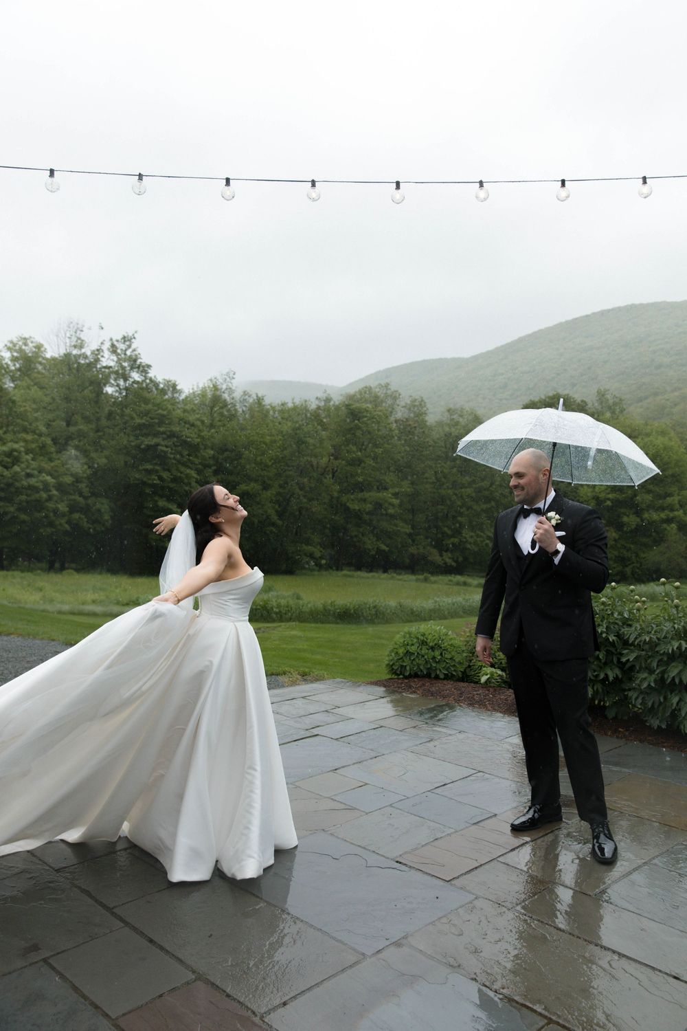 Newlyweds dance and twirl playfully with umbrellas in the rain on a stone patio surrounded by misty green hills.