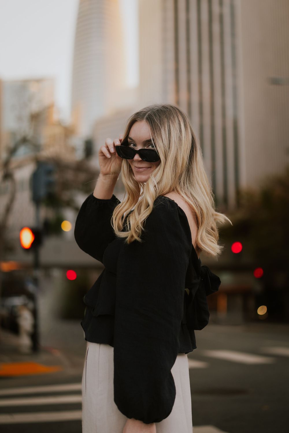 Blonde woman in black jacket and sunglasses posing on urban street with city buildings in background.