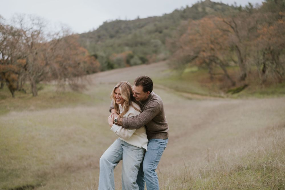 Couple laughing and embracing in a scenic meadow with rolling hills and autumn trees in the background.