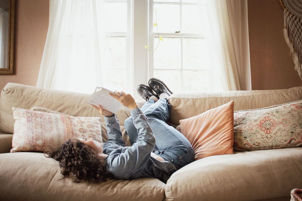 Person lying on beige couch reading book in sunlit room with decorative pillows.