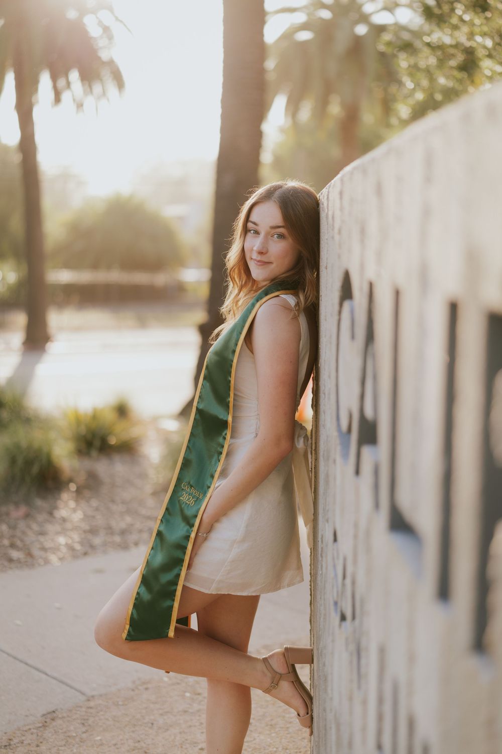 Young woman in white dress and green graduation sash leans against a wall outdoors in golden hour light.