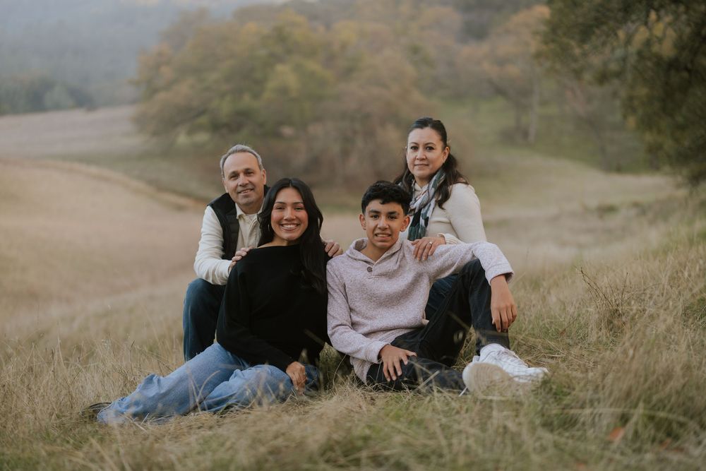Family of four smiling together outdoors on a grassy hillside with autumn scenery in the background.