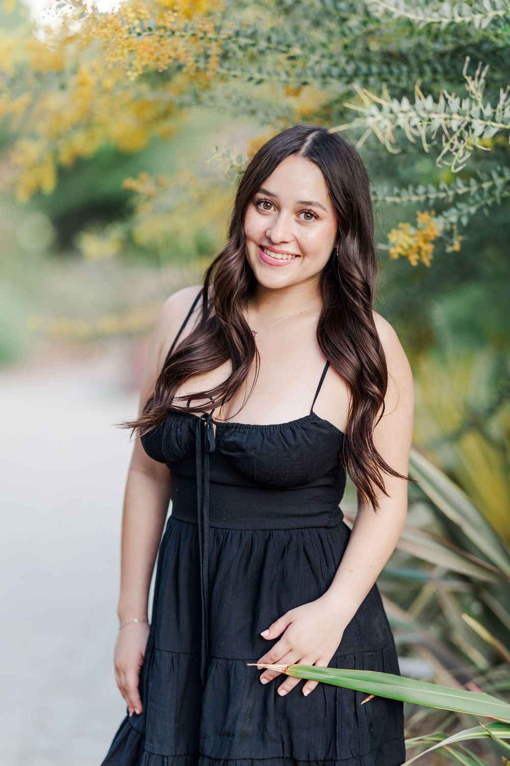 A portrait surrounded by yellow flowering plants and greenery in a black dress on a garden path.