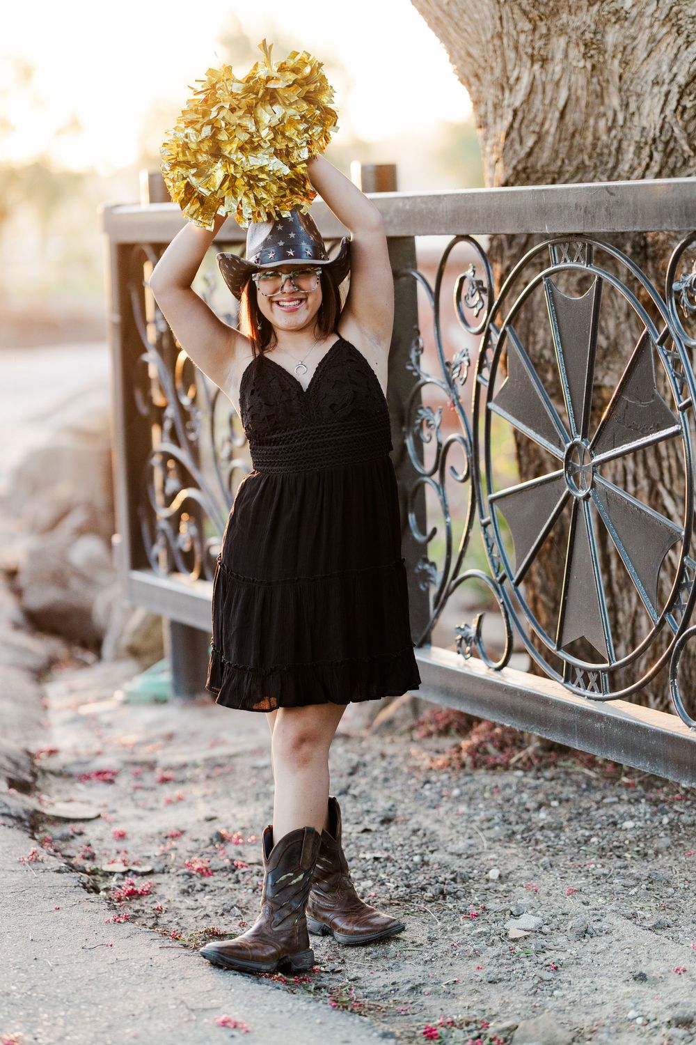 A cheerleader in a black dress and cowboy boots holds up gold pom poms next to a decorative metal railing at sunset.