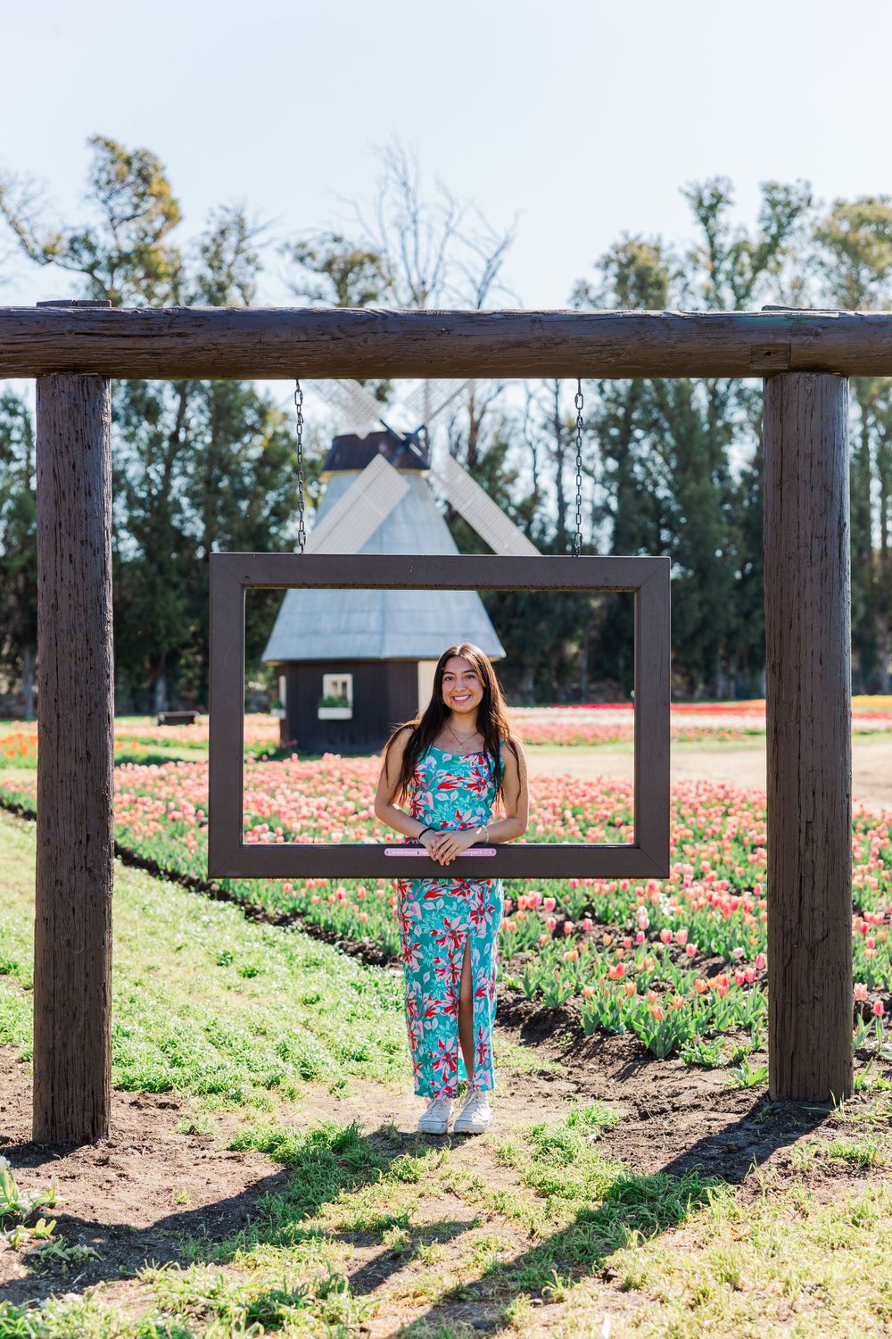 Person in floral jumpsuit posing under wooden frame structure with windmill in background.