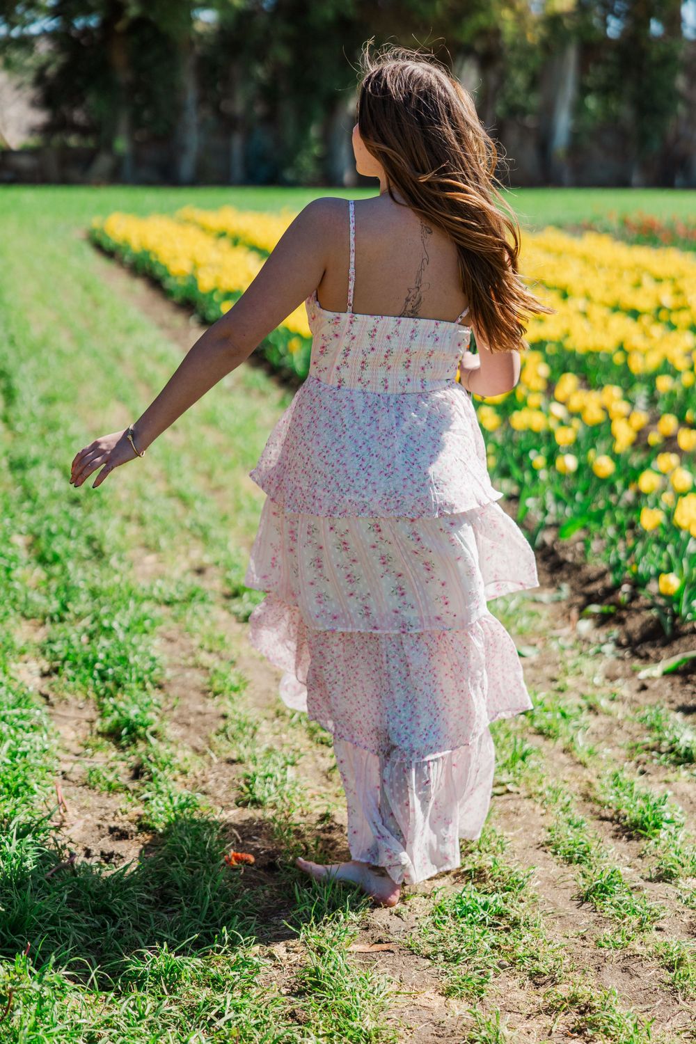 Back view of woman in white tiered sundress walking through field of blooming yellow daffodils on sunny spring day.