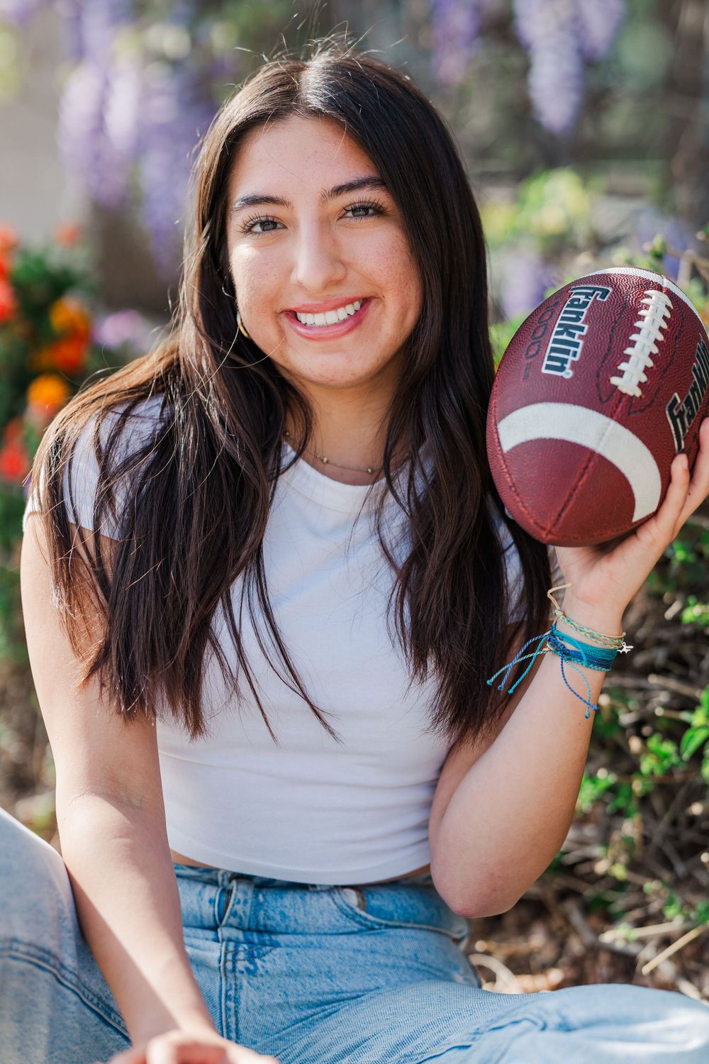 Person in white top and jeans holding a football poses outdoors with flowers in the background.