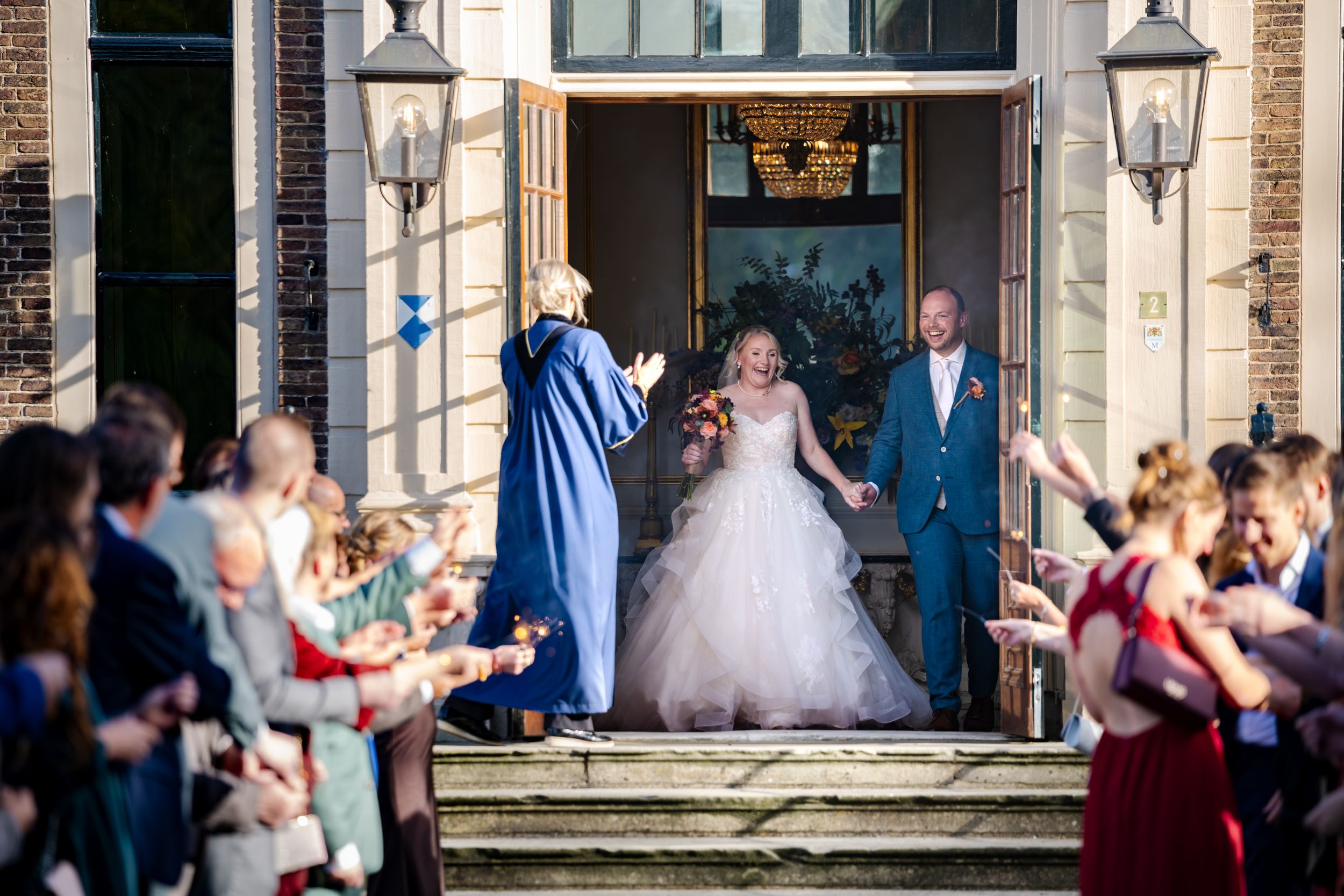 Blij bruidspaar loopt naar buiten na hun trouwceremonie in kasteel Ruurlo en worden opgewacht door hun trouwgasten met sterretjes.