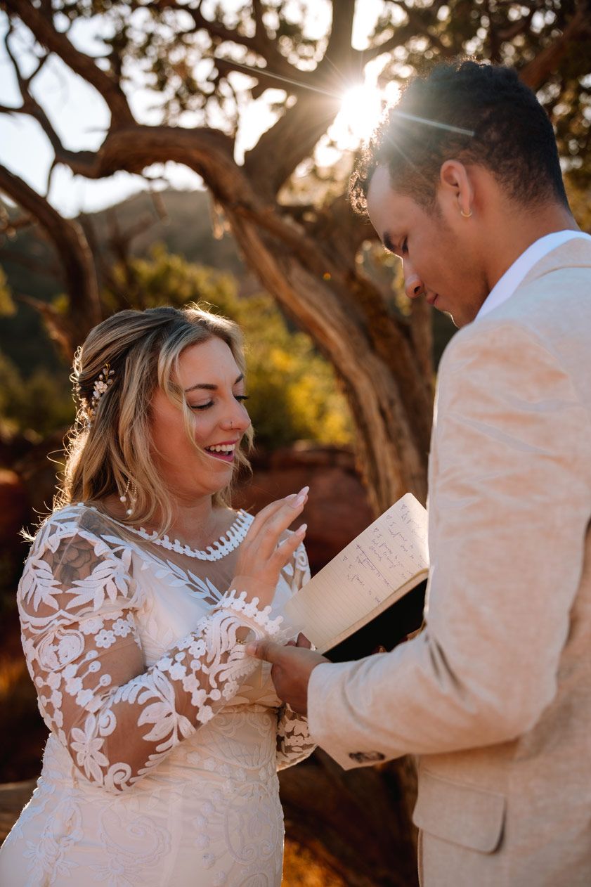 A joyful wedding moment during vow reading in a Sedona desert setting with sun flare through trees.