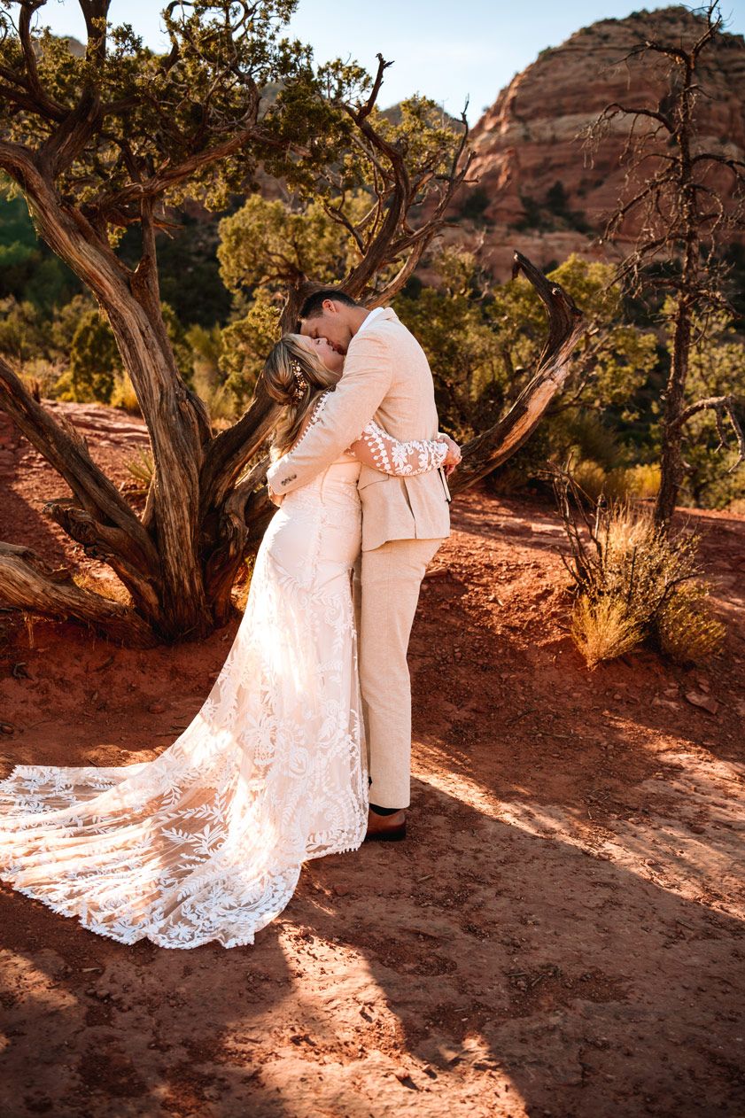A wedding couple embraces under a twisted tree in a dramatic Sedona red rock desert landscape.