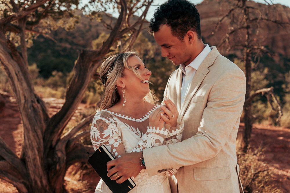 Wedding couple embraces against Sedona red rock desert landscape during sunset, wearing white dress and beige suit.