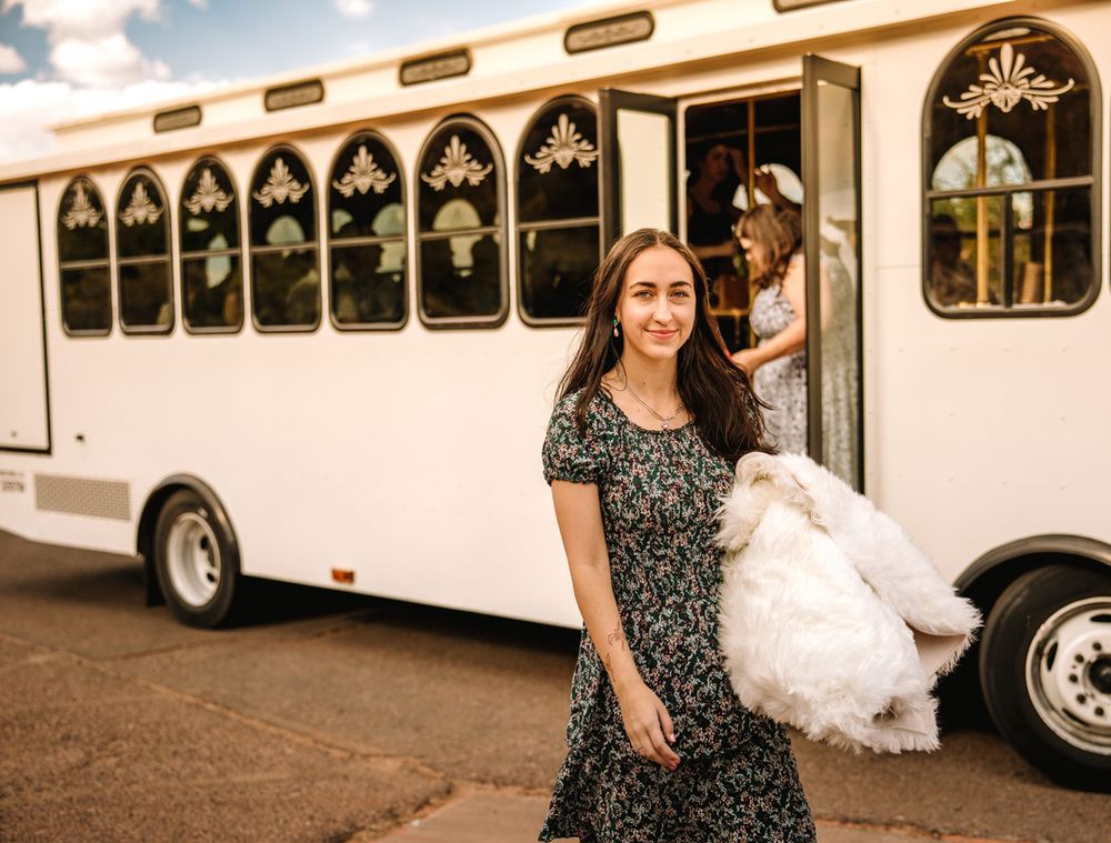 A stylish wedding party member exits a white trolley bus carrying formal attire.