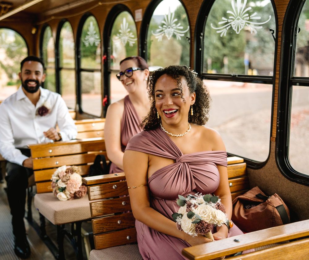 Wedding party members seated inside a vintage trolley car with wooden benches and arched windows.