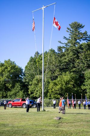 Flag poles with American and Canadian flags standing tall against blue sky with spectators below.