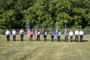 Line of people holding American and other flags walk across a grassy field during an outdoor event.