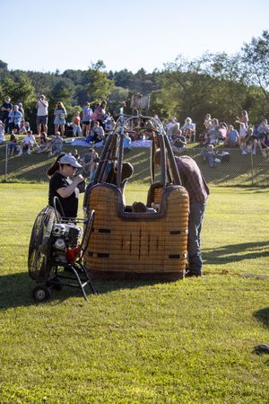 Wicker balloon baskets lined up on a grassy hill with crowds gathering around.