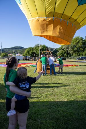 People gather around a yellow hot air balloon being inflated in a grassy field with mountains in the background.