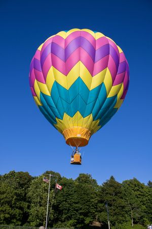 A vibrant rainbow-colored hot air balloon rises against a deep blue sky with trees in the background.