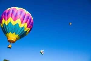 Multicolored hot air balloons soar through a clear blue sky during a summer festival.