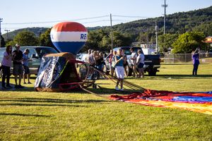 Hot air balloon festival setup with equipment and crowds spread across a sunny field near mountains.